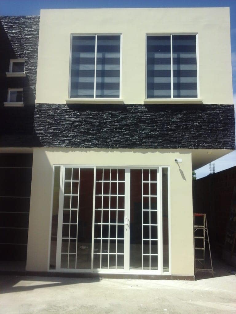 The exterior of a two-story building features a modern design with a contrasting stone facade. Large, striped windows allow natural light into the upper level. French doors at the entrance add an inviting touch, while a staircase is visible to the side.