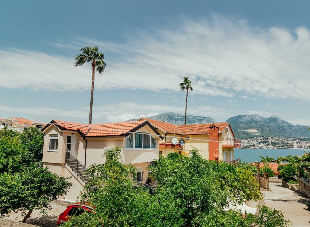 A two-story building is framed by palm trees and greenery, with red-tiled roofs contrasting against a clear blue sky. A glimpse of the water and mountains is visible in the background, emphasizing the peaceful setting of the studio apartment.