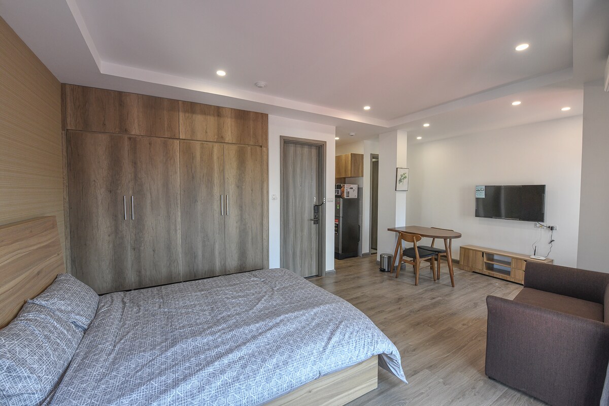 A modern studio apartment features a spacious bed with patterned bedding, adjacent to a compact dining area with a wooden table and chairs. A brown armchair sits nearby, while a wall-mounted TV is positioned across from the bed. An equipped kitchenette is visible in the background.