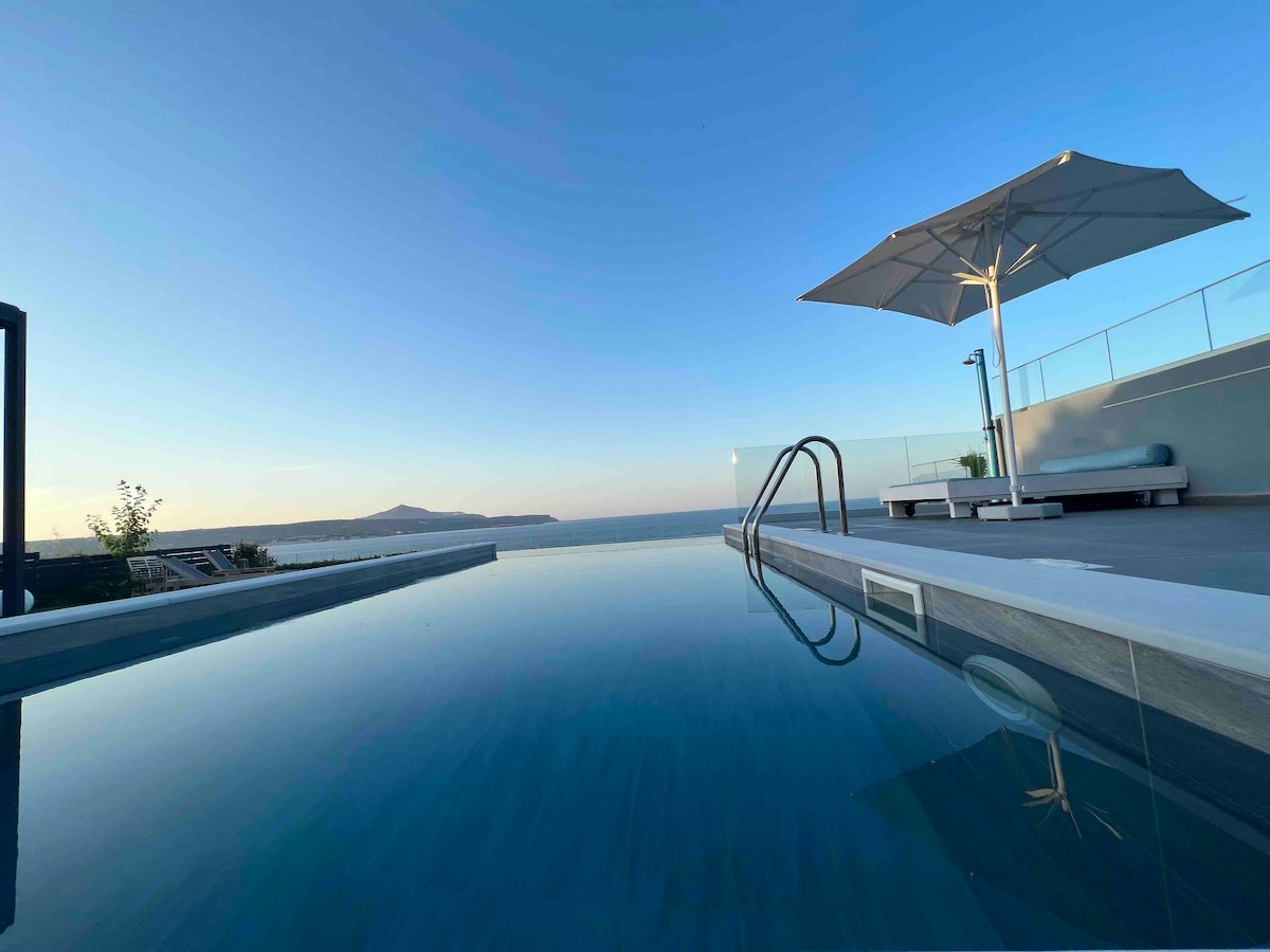 An expansive infinity pool reflects the clear sky, seamlessly blending with the horizon. A large, shaded umbrella and lounging area are positioned nearby, providing a serene space for relaxation. The scene captures a tranquil coastal view, enhancing the villa's inviting atmosphere.