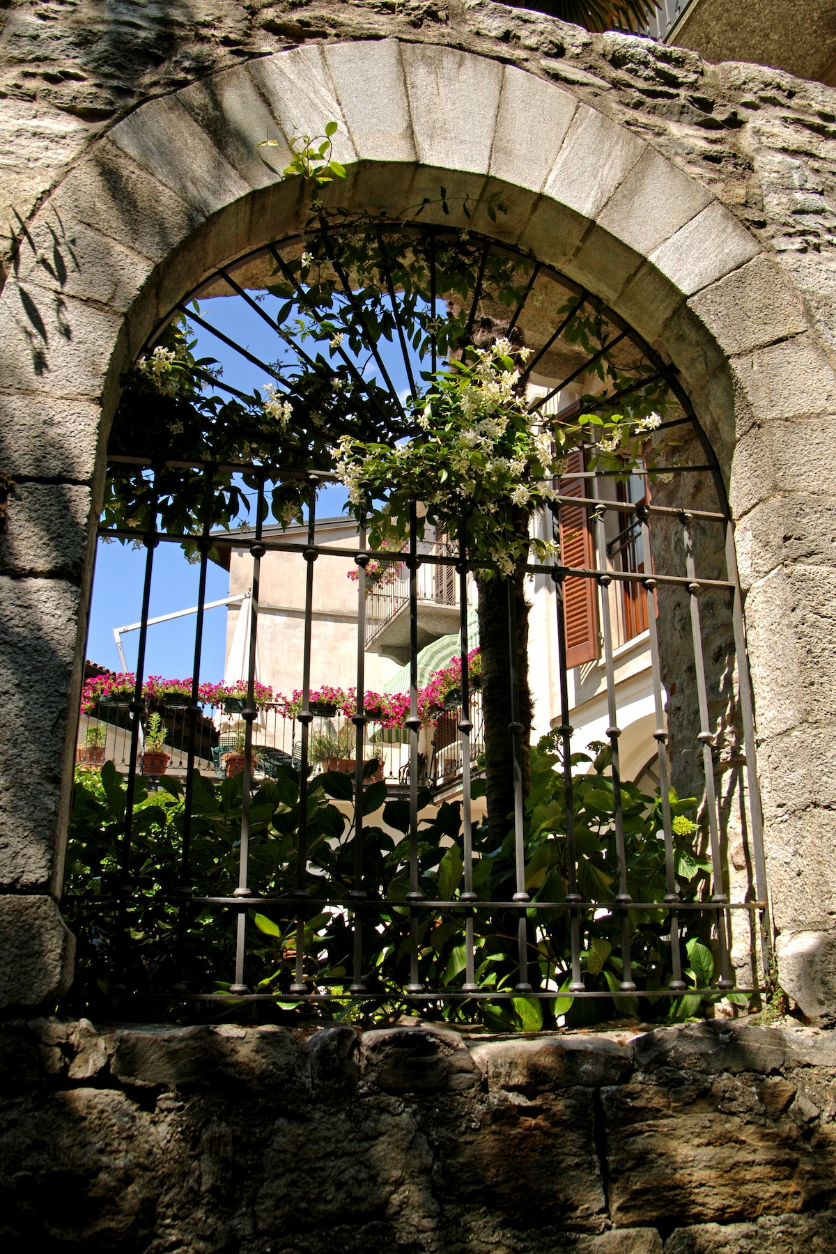 An archway framed by stone features a wrought iron gate, allowing glimpses of greenery and blooming flowers. Light filters through the openings, highlighting the natural elements within the courtyard beyond.