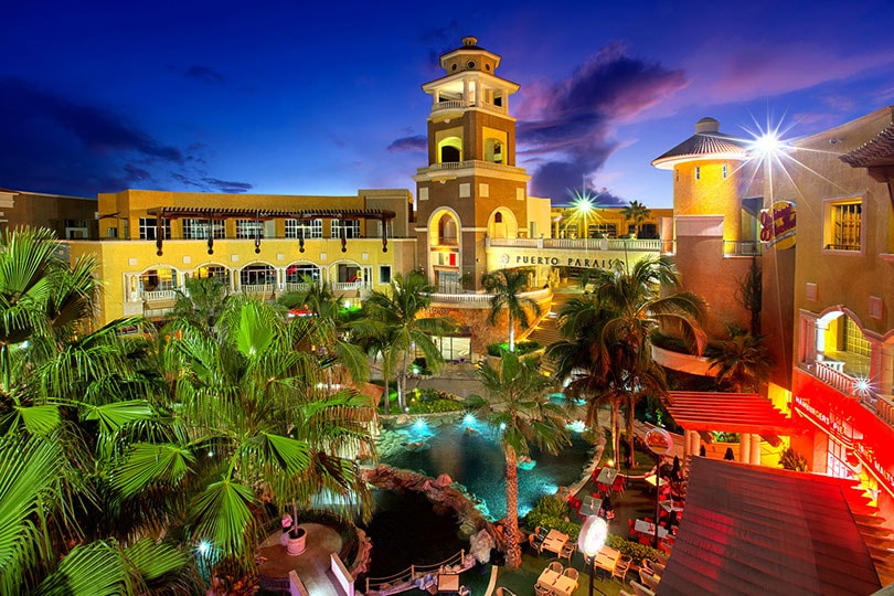 The image captures a vibrant evening view of Puerto Paraiso Mall, featuring lush palm trees surrounding a serene pool area. Warm lighting illuminates the architecture, highlighting the mall's distinctive design. A mix of outdoor seating and water features adds to the inviting ambiance.