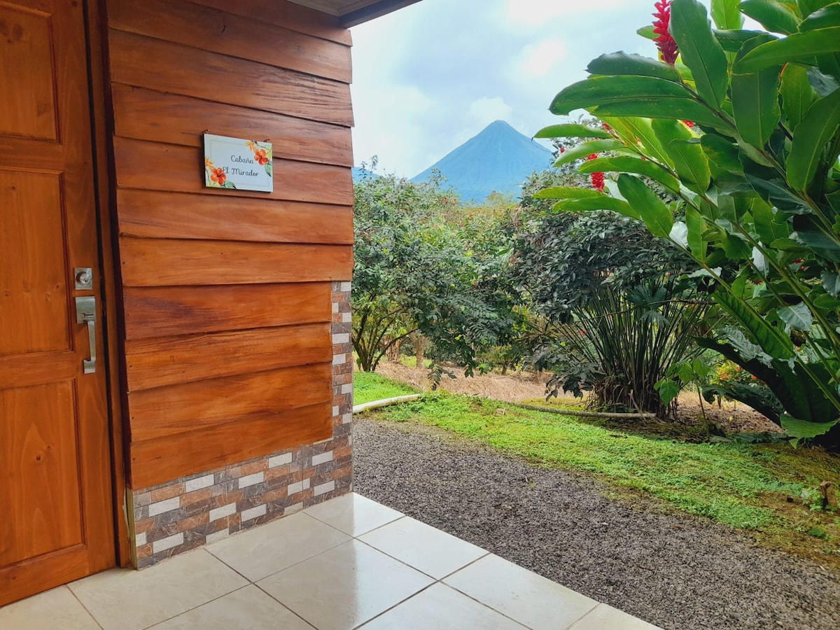 The entrance of the cabin features wooden paneling and a welcoming sign. Lush green plants surround the doorway, with a view of Arenal Volcano in the background. A stone pathway leads to the entrance, highlighting the serene rural setting.