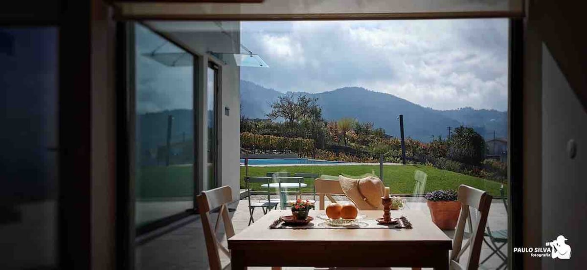 A dining table is set with fruit and minimalist decor, positioned near expansive glass doors that open to a scenic view of the hills and vineyards. A swimming pool is visible in the distance, enhancing the connection to the natural surroundings.