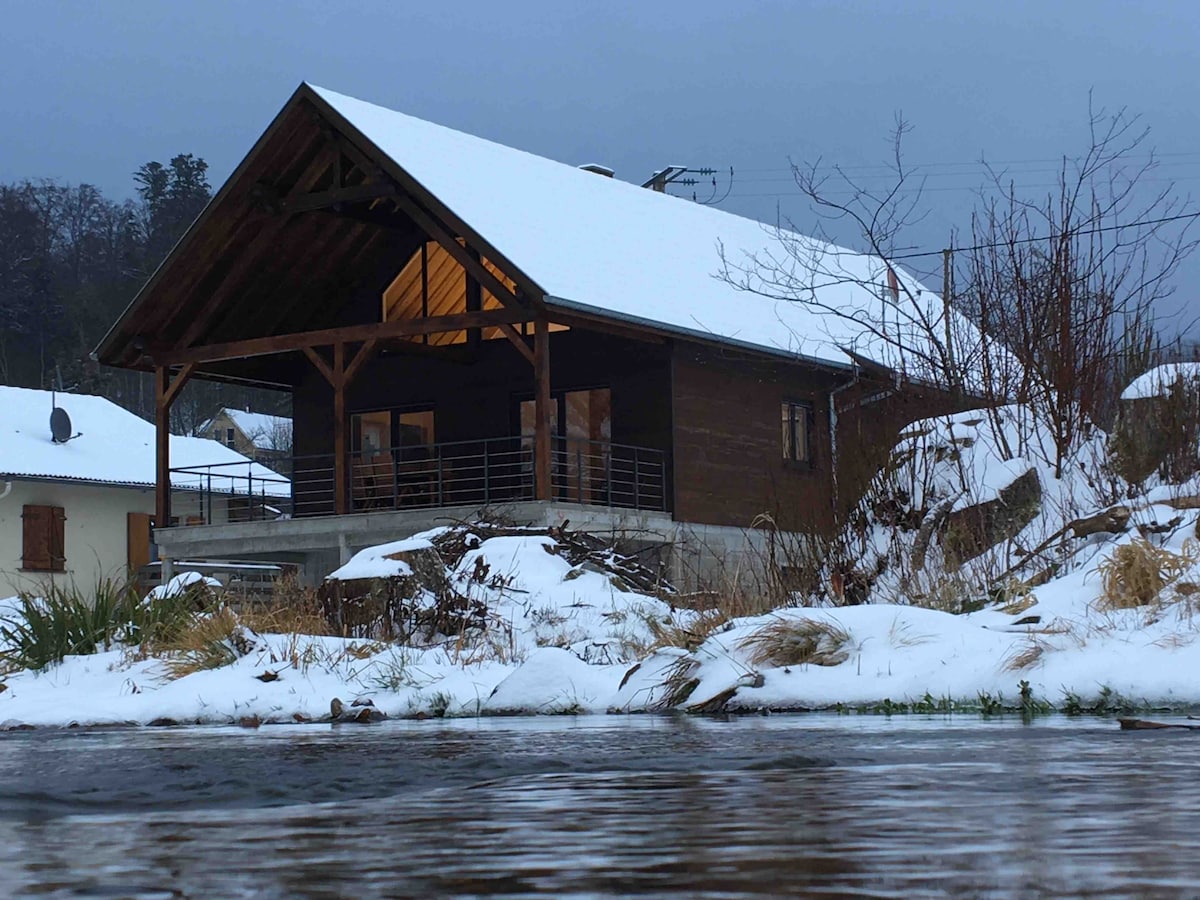 A modern chalet with a sloped roof is framed by a snowy landscape. A spacious terrace overlooks the gentle river, reflecting the soft light of dawn. Surrounding vegetation is dusted with snow, contributing to a serene winter ambiance.