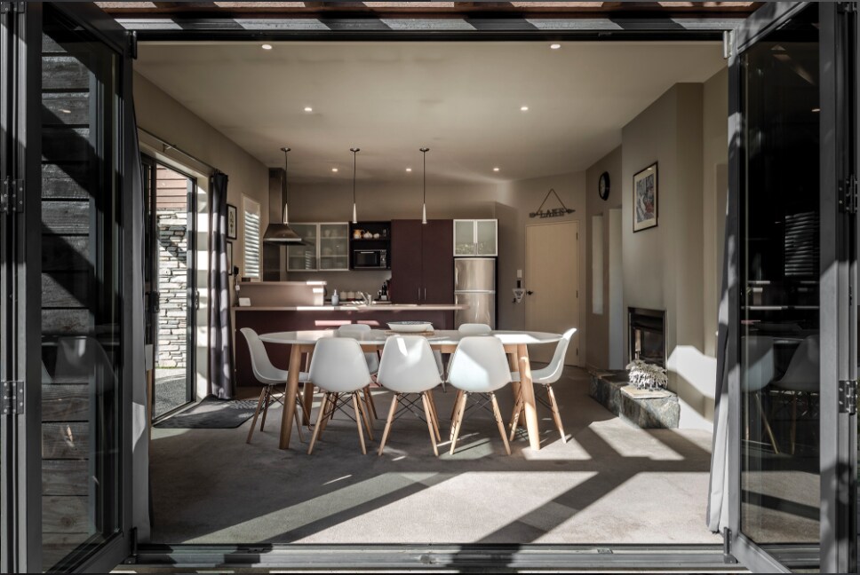 An open-plan dining area is visible through bi-fold doors, featuring a large table surrounded by white chairs. A modern kitchen with stainless steel appliances is positioned in the background, alongside a cozy sitting area with a gas fireplace.