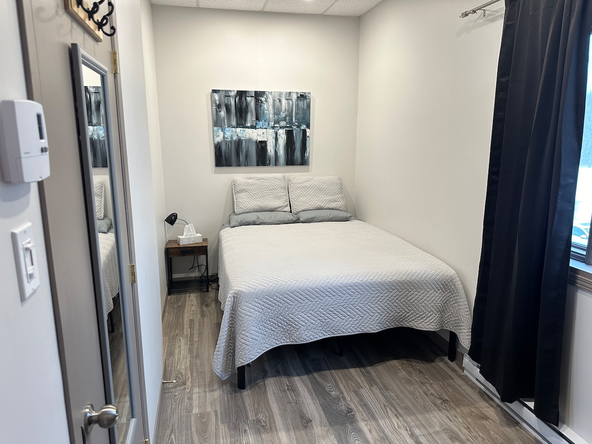 A simple sleeping area features a spacious bed covered with a light quilt. A modern piece of wall art hangs above the bed. Natural light enters through a window, dressed with dark curtains, while a nightstand and lamp are positioned beside the bed.