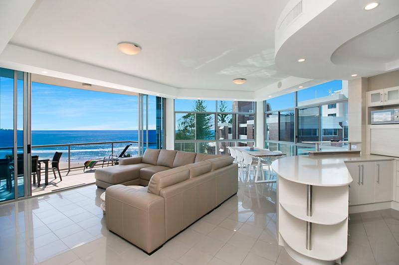 An open living area features a light-colored sectional sofa facing large windows with ocean views. A modern kitchen is situated nearby, showcasing a curved counter and well-defined spaces. The outdoor patio is visible through glass doors, offering additional seating and stunning coastal scenery.