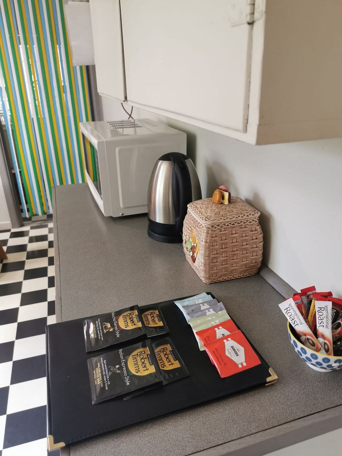 The kitchen countertop is arranged with a kettle, a microwave, and a woven basket. A selection of tea and coffee varieties is displayed on a black placemat, along with various pantry condiments. The checkerboard flooring and colorful striped curtains add a lively touch.