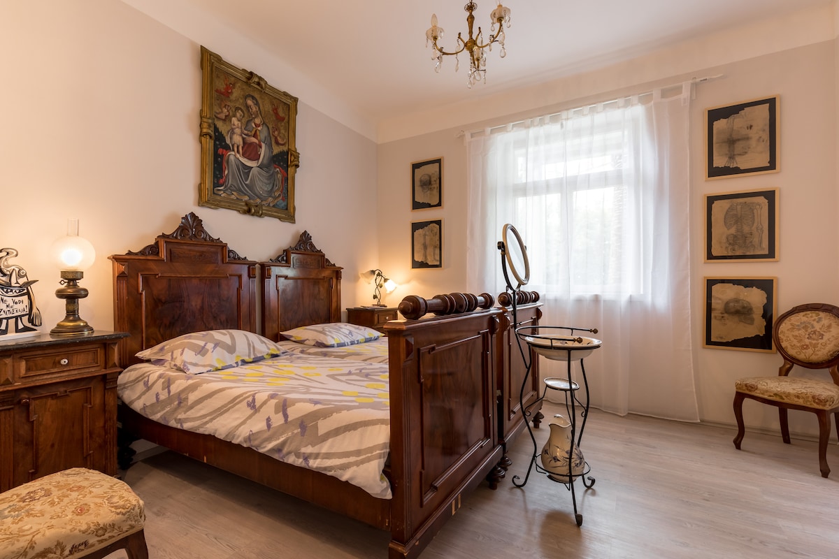 A warmly styled bedroom features a wooden bed with patterned bedding and accent pillows. Soft light filters through a sheer curtain, illuminating an antique dresser and a small vanity table. Elegant artwork adorns the walls, enhancing the room's historical charm.