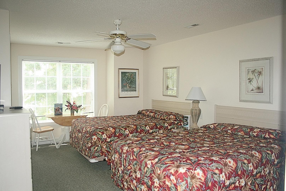A spacious room features two beautifully patterned beds with decorative bedspreads. A large window allows natural light to fill the space. A small dining table with two chairs and a lamp is situated nearby, complemented by framed artwork on the walls.