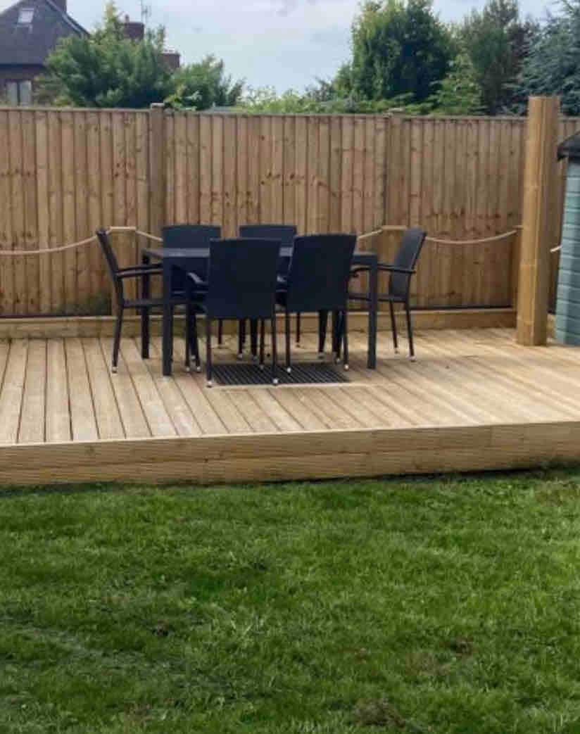 A wooden deck sits atop a well-maintained lawn, featuring a rectangular table surrounded by six black chairs. The area is enclosed by a wooden fence, creating a private and inviting outdoor space.