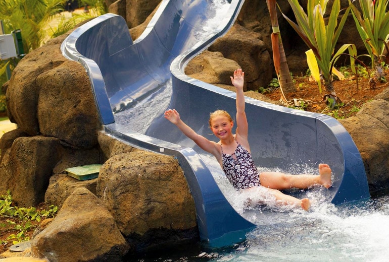 A sleek water slide curves down amid natural rock formations and lush greenery. Water cascades from the slide, creating splashes as a child navigates downwards with raised arms, showcasing a playful atmosphere designed for family enjoyment.