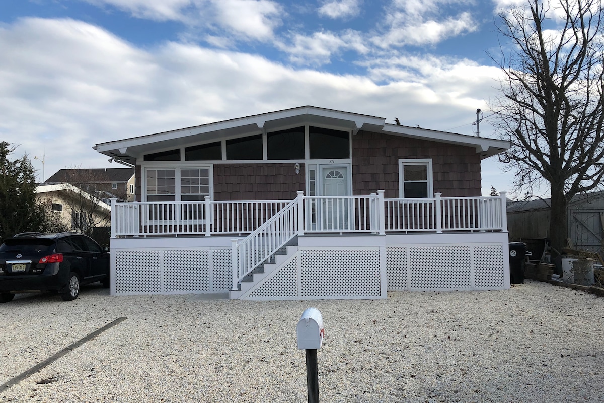 The exterior of a spacious waterfront home is displayed, featuring a well-maintained porch with white railing and steps leading to the entrance. Gravel surrounds the property, providing a clean and organized appearance. Windows reflect natural light against a partly cloudy sky.