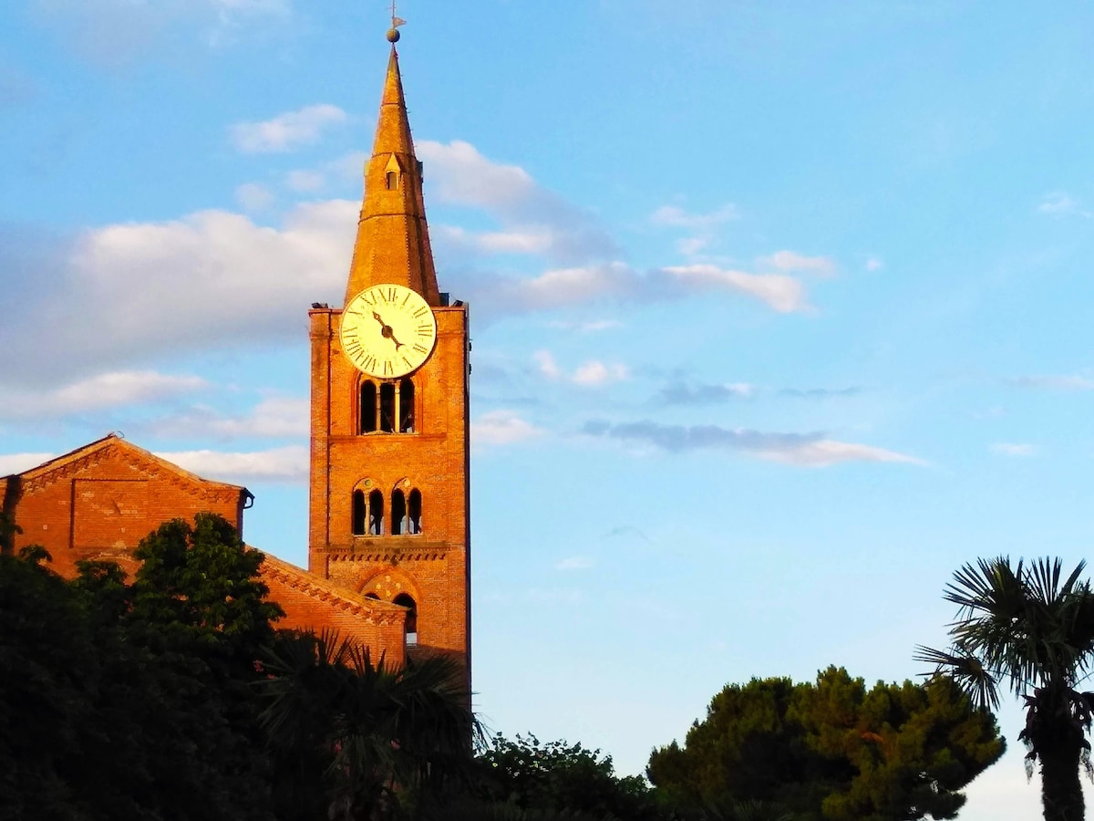 A tall brick clock tower rises against a clear sky, showcasing intricate architectural details. The clock face, visible at the top, is surrounded by soft evening light. Lush greenery and palm trees frame the scene, adding a touch of nature to the historic structure.