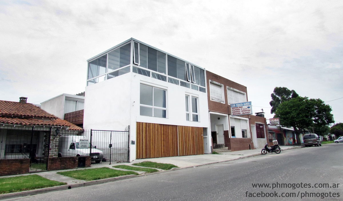 A modern two-story building is presented with a clean, minimalist façade featuring large windows and wooden accents. The design emphasizes natural light and openness. Surrounding areas are landscaped, complementing the structure's contemporary style.