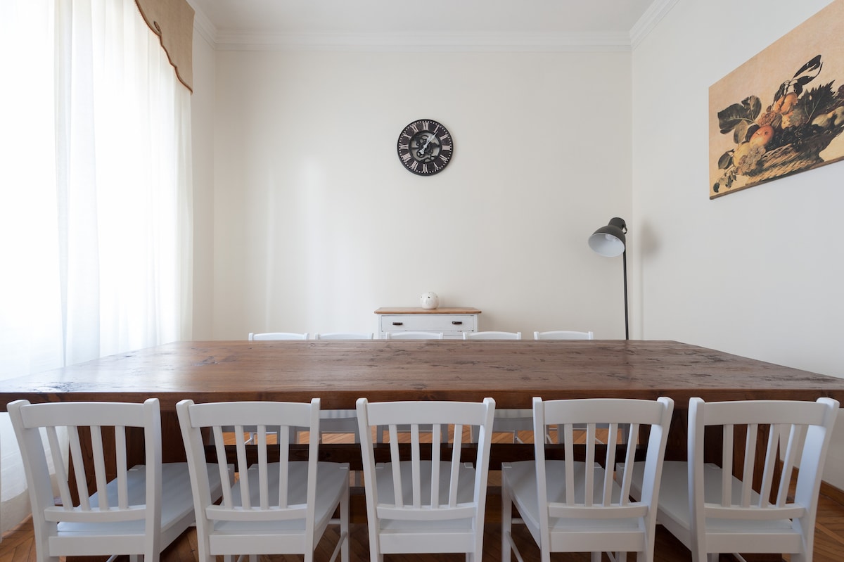 A spacious dining area is presented with a large wooden table, surrounded by six white chairs. A wall clock adds functionality, while a piece of artwork decorates one side. Natural light flows in through large windows, enhancing the warmth of the room.