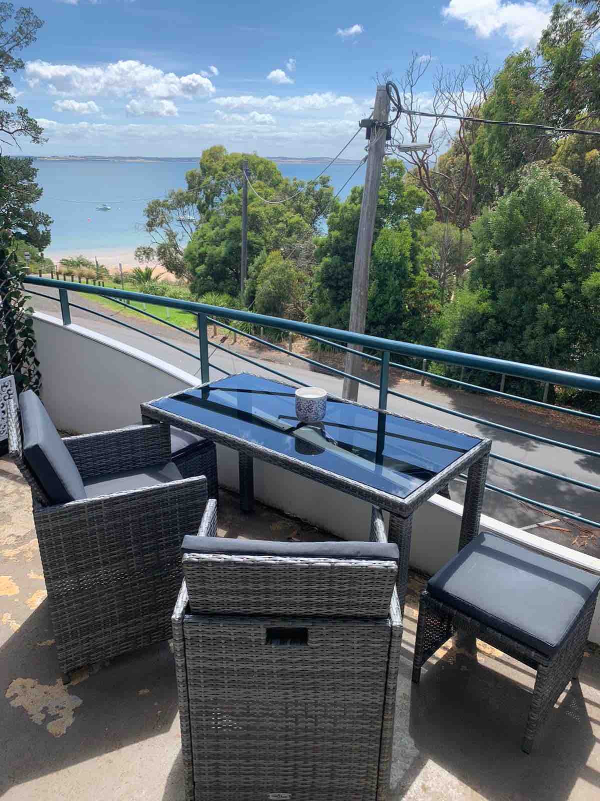 A balcony seating area features a glass-top table surrounded by four rattan chairs. Views of the coastline and trees are visible in the background, while the ocean extends beyond the land. The scene is set under a clear blue sky with scattered clouds.