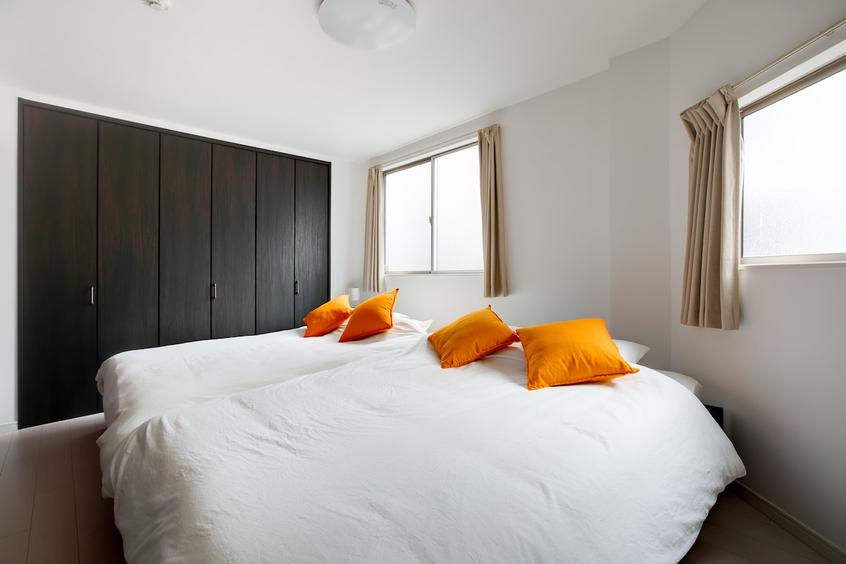 The room features two spacious beds dressed in white linens, complemented by bright orange pillows. Natural light enters through two windows, enhancing the clean and modern decor. A dark wooden wardrobe provides storage, while beige curtains frame the windows.