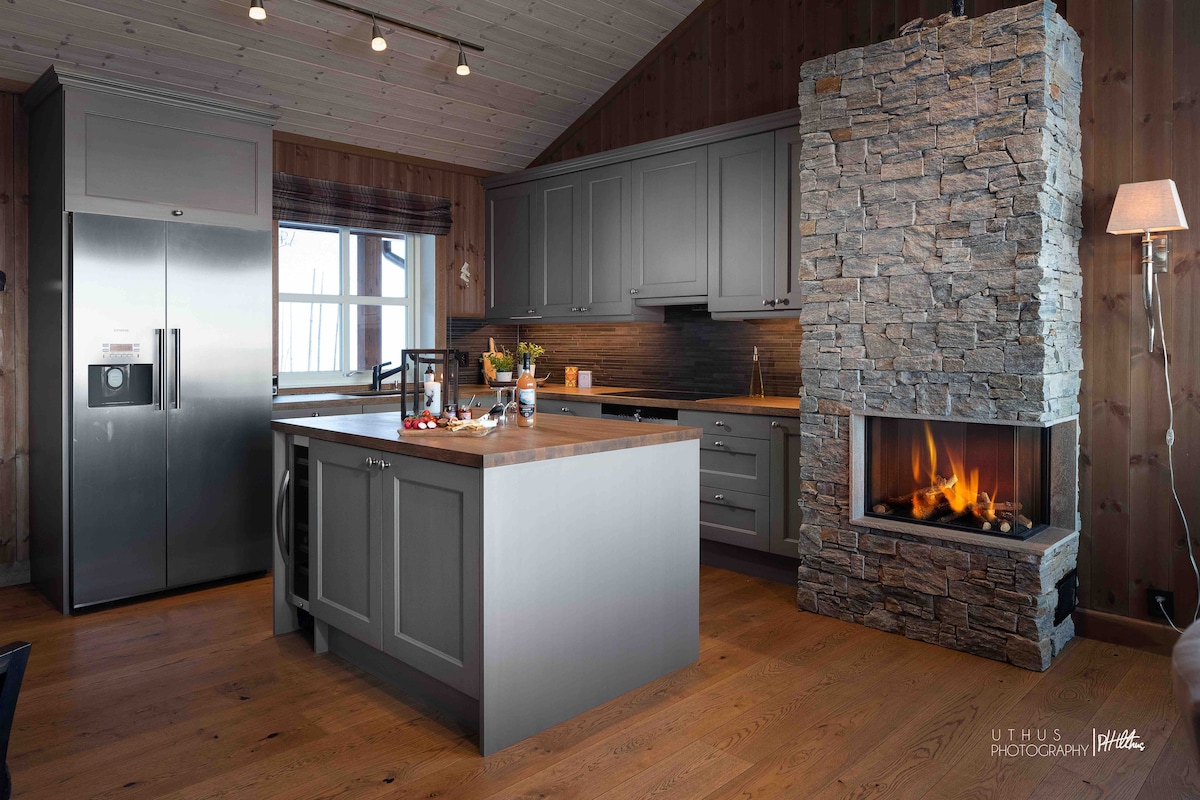 A modern kitchen is presented with grey cabinetry and a large island. Stainless steel appliances, including a refrigerator, complement the stone fireplace that adds warmth. Natural light streams through the window, illuminating the wooden walls and floor, creating a welcoming space for meal preparation.