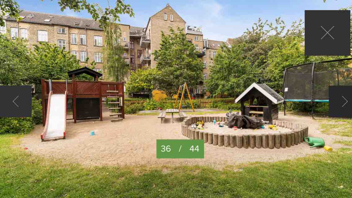 A family-friendly backyard playground is shown, featuring a wooden climbing structure with a slide, swings, and a sandbox surrounded by logs. A trampoline and a small playhouse are visible, along with various toys scattered in the sandy area and lush greenery in the background.