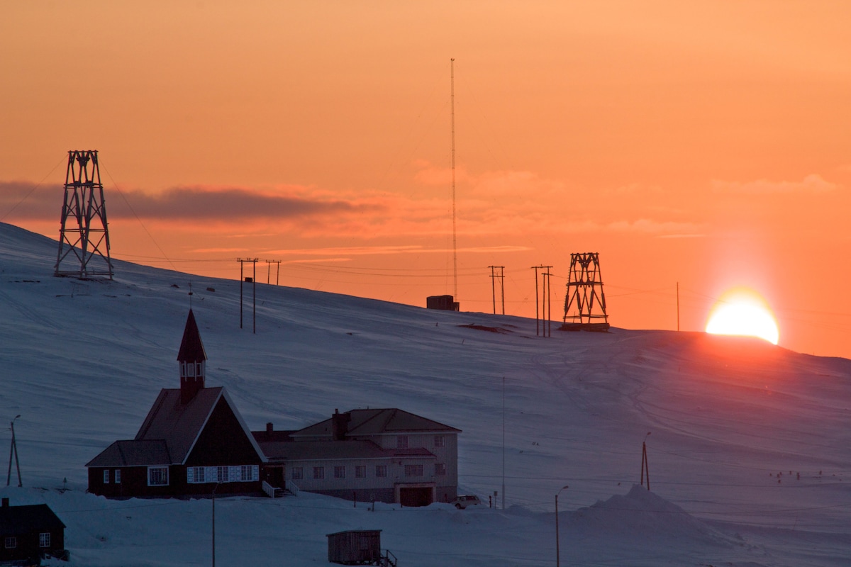 The image captures a serene winter landscape at sunset, with the sun dipping below the horizon. Silhouettes of wooden structures and power lines are visible against the snow-covered terrain, while a church spire protrudes from the lower foreground, contributing to the tranquil atmosphere.