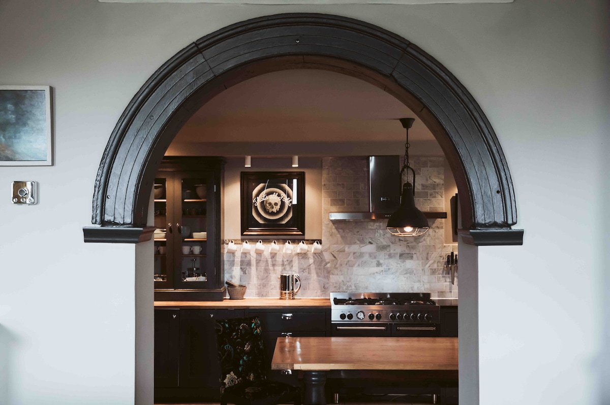 An elegant kitchen is framed by an arched entryway, showcasing dark cabinetry and modern appliances. The space features a rustic wooden dining table accompanied by a stylish pendant light. The backsplash is adorned with subtle stone tiles, enhancing the design's visual appeal while offering functionality.