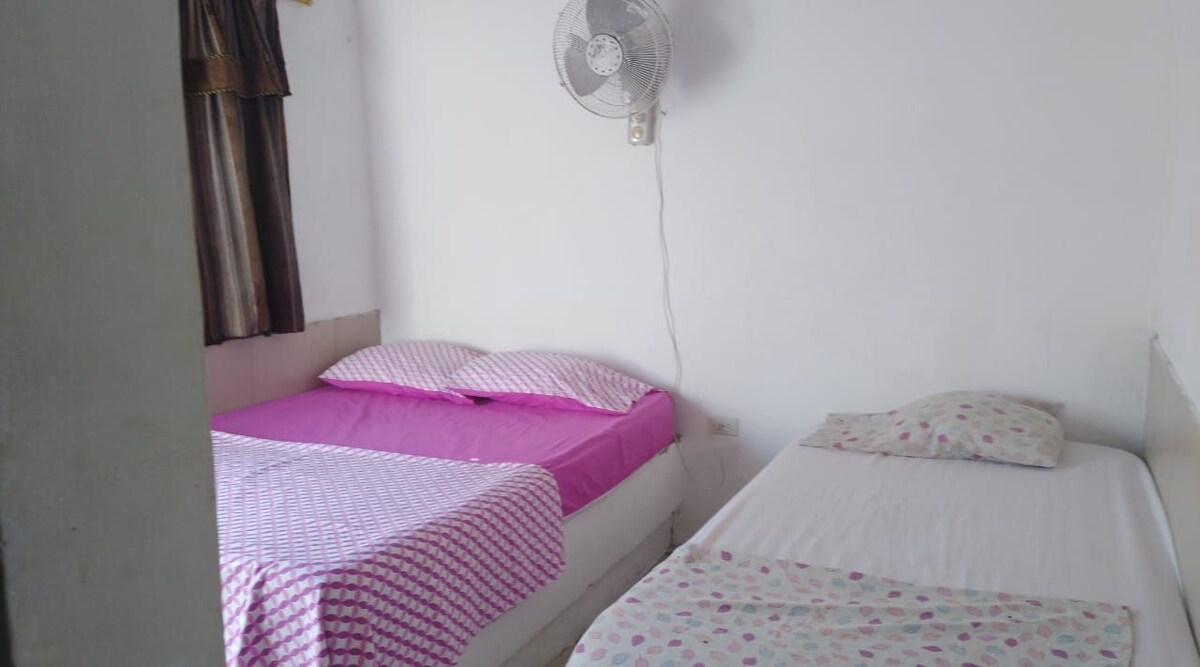A simple sleeping area is presented with two beds, one featuring pink sheets and the other covered with a white sheet adorned with polka dots. A ceiling fan hangs above to provide airflow, and natural light enters through a nearby window with dark curtains.