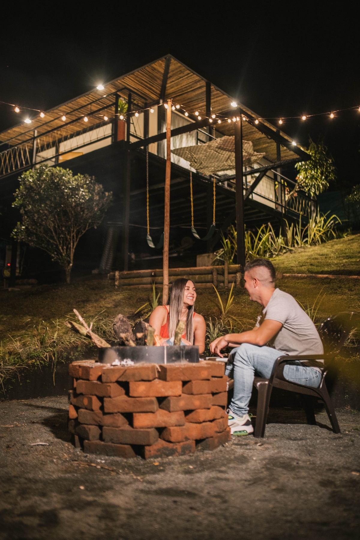 A well-lit outdoor area features a brick fire pit surrounded by seating. In the background, a two-story cabin is illuminated by string lights, providing a cozy ambiance. Lush greenery surrounds the space, contributing to a serene outdoor environment.