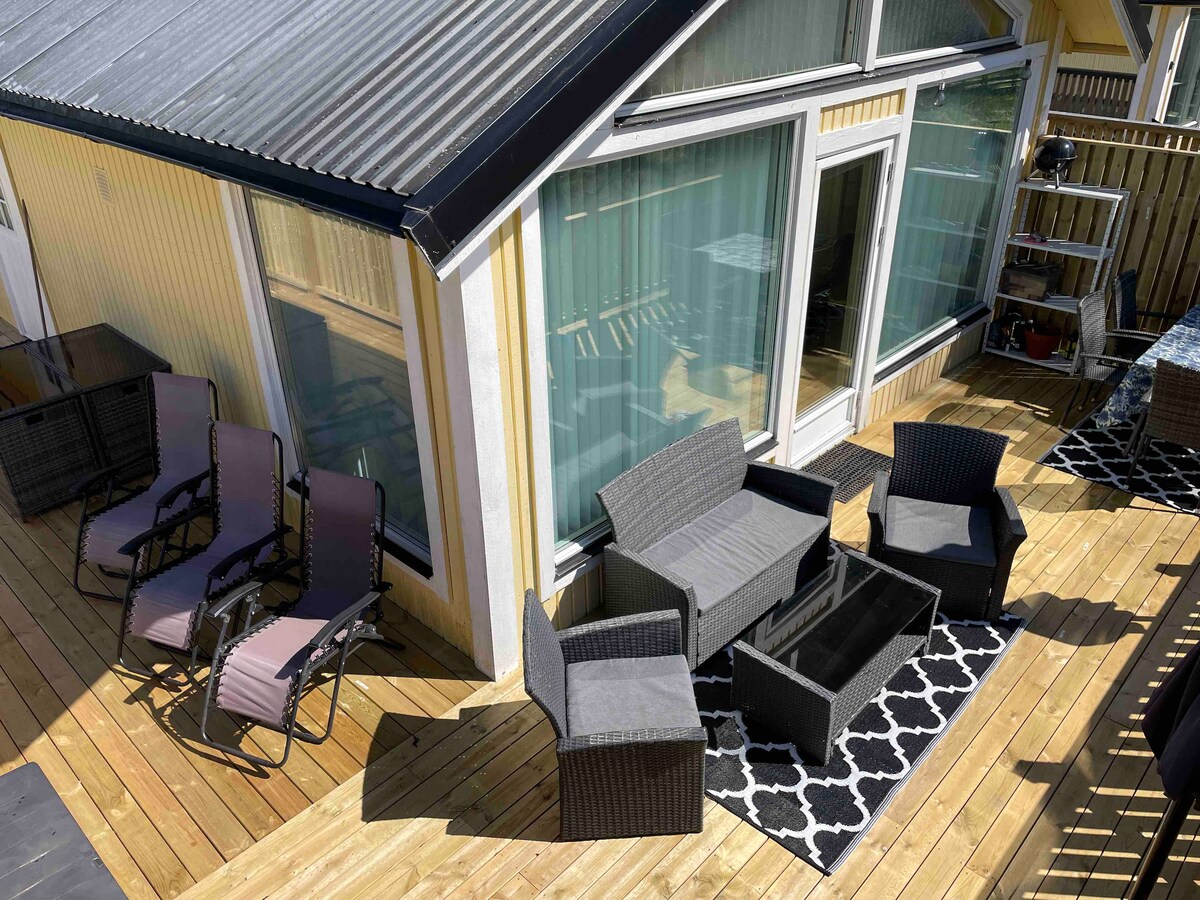 The spacious wooden deck is furnished with several comfortable seating options, including two loungers and a set of armchairs surrounding a central coffee table. Large glass doors allow natural light to fill the area, enhancing the connection with the outdoors.