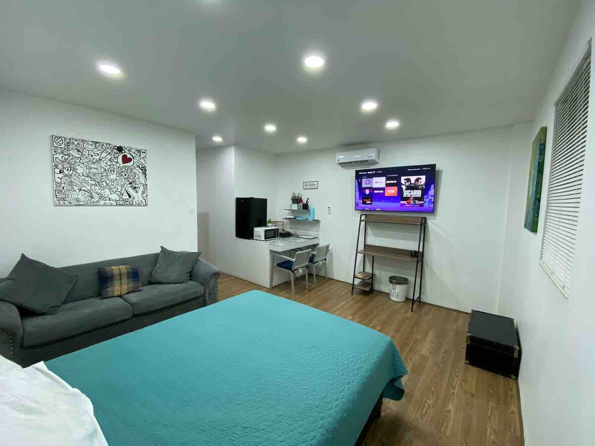 A spacious room is presented with a large, turquoise bedspread covering a wide bed. A cozy couch sits to one side, while a mounted television is displayed on the opposite wall. A small kitchenette is visible in the corner, providing essential amenities.