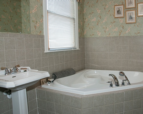 A bathroom is depicted featuring a spa tub and a pedestal sink. The tub is positioned near a window adorned with blinds, allowing daylight to brighten the space. Floral wallpaper adds a soft touch to the tiled walls, contributing to the overall design.