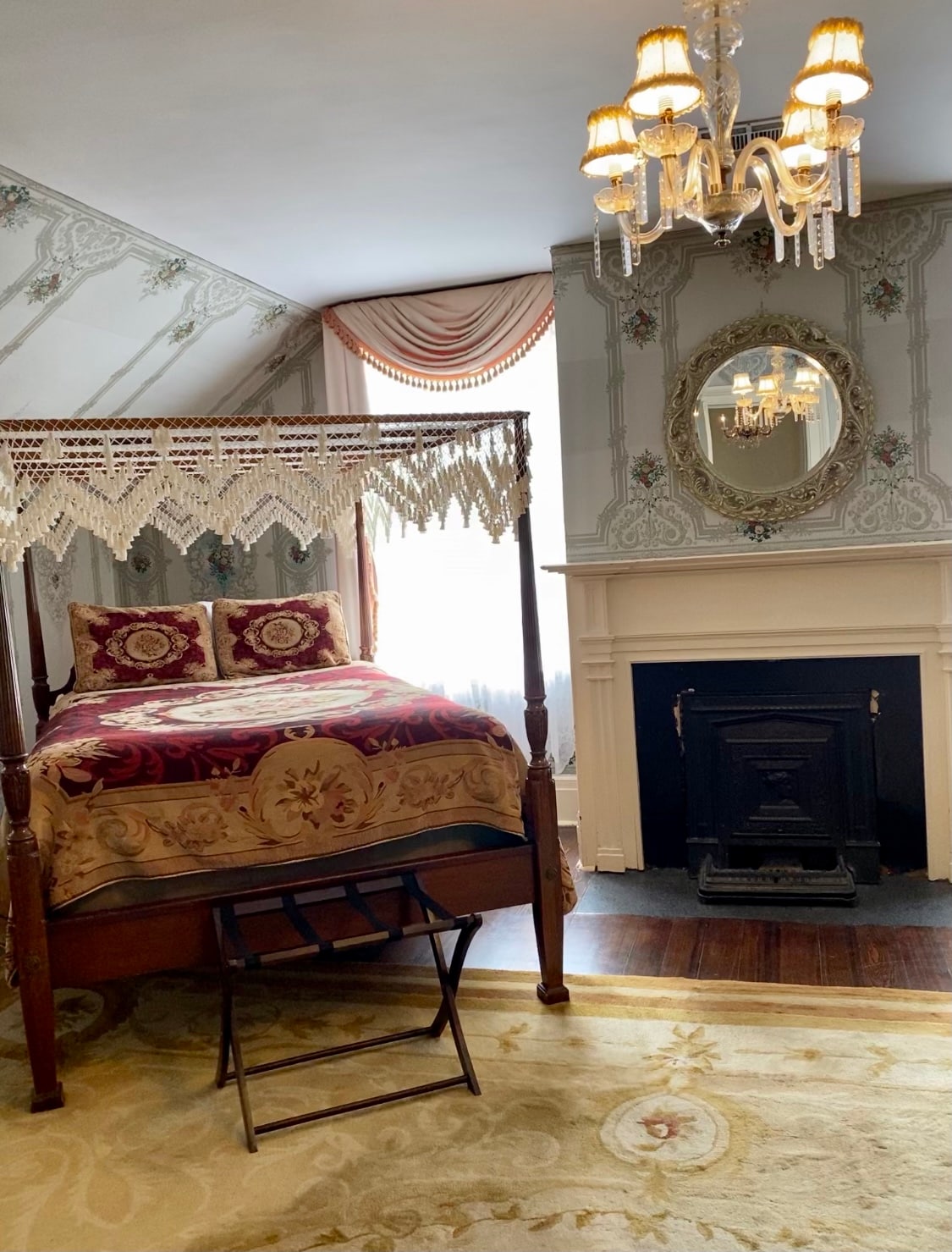 A spacious room features a queen size four-poster bed adorned with decorative bedding. An antique fireplace is positioned beneath a mirror, with a chandelier overhead providing soft lighting. Elegant wallpaper and large windows create a serene atmosphere.