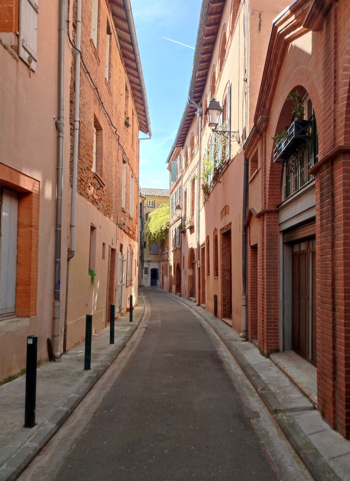 A narrow lane lined with charming buildings featuring warm-colored facades. Distinctive brick accents are evident, along with greenery draping from a balcony overhead. Street lamps and black bollards add functional elements to the environment, while the bright blue sky is visible above the rooftops.