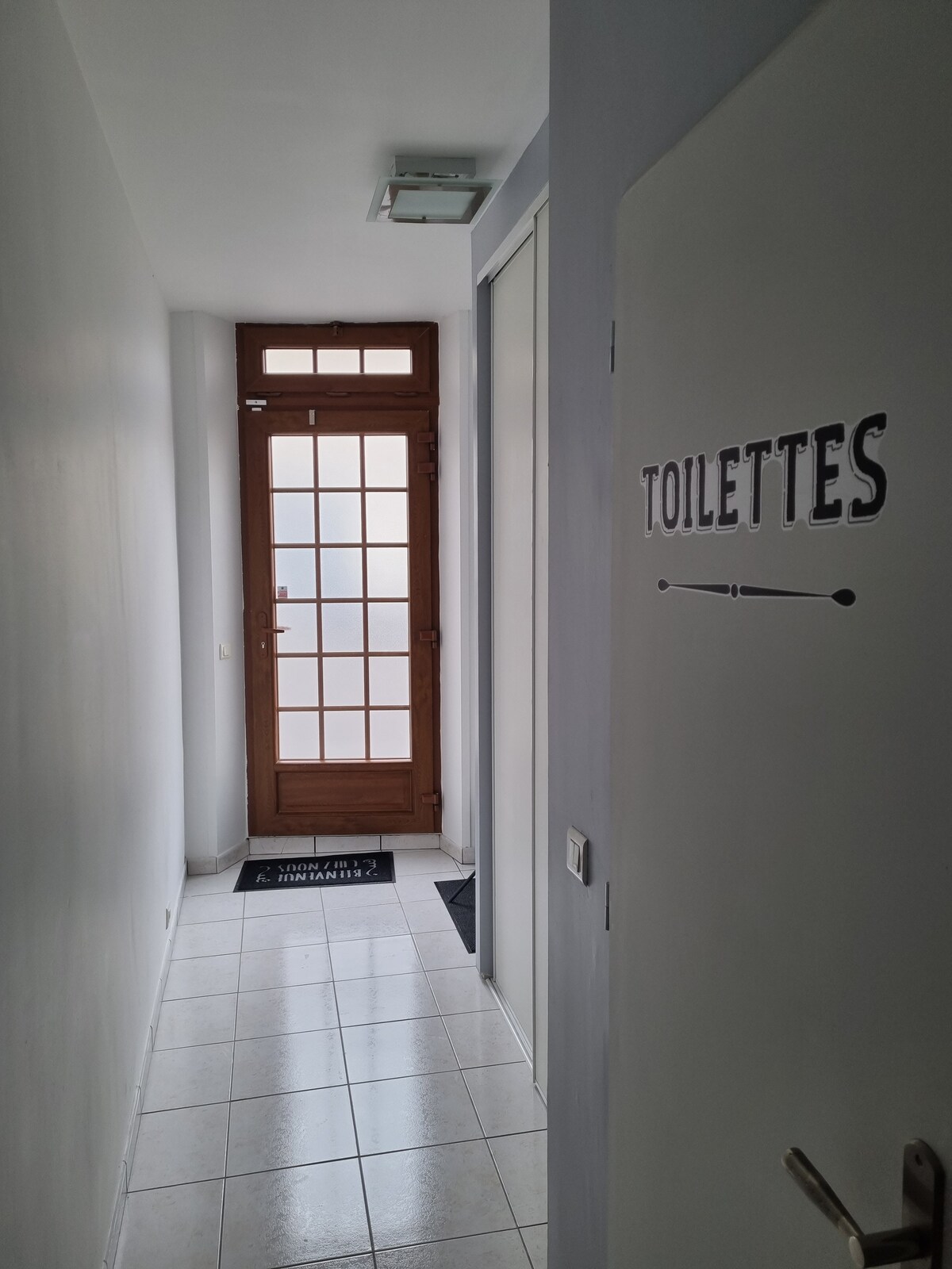 A hallway is visible, featuring a door marked 'TOILETTES' on the right. Light enters through a glass-paneled main entrance door at the end of the corridor, which is characterized by white walls and tiled flooring.