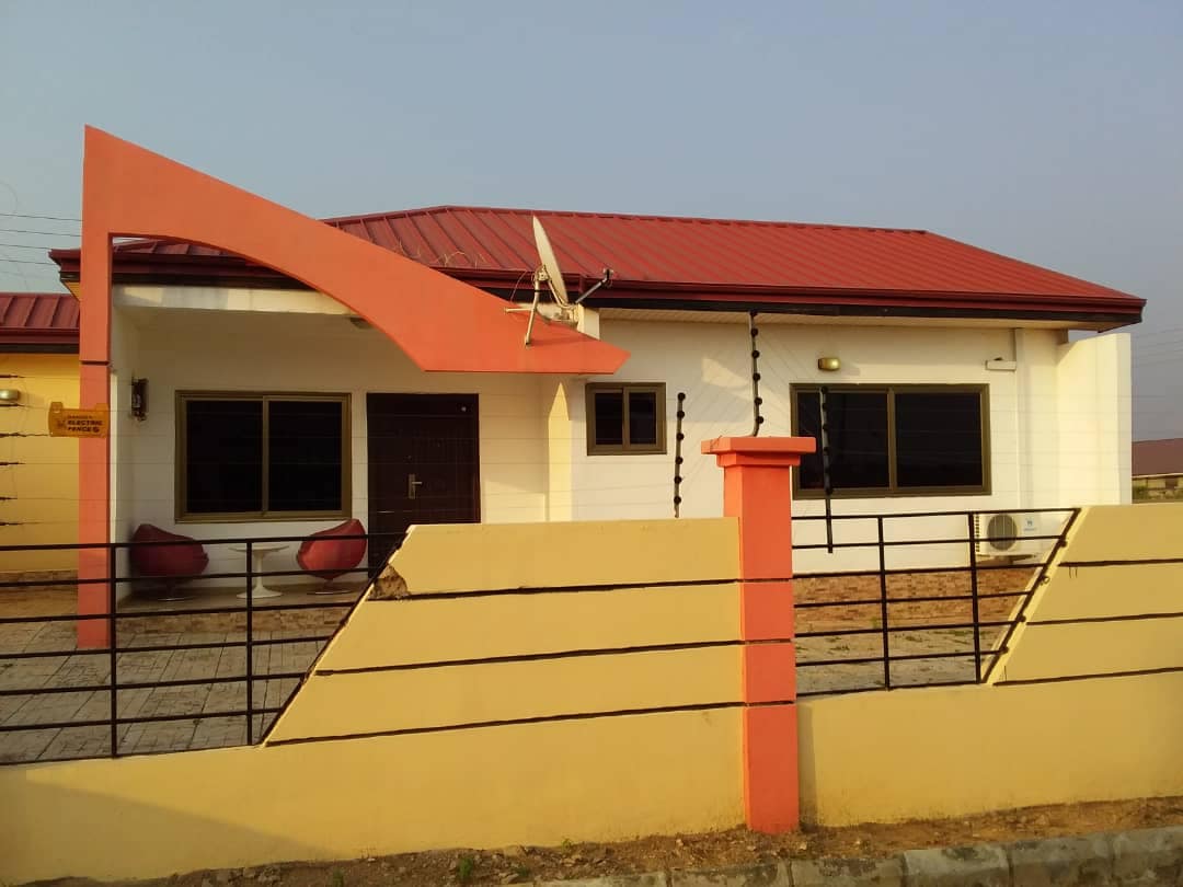 The exterior of a single-story home features a sloped red roof and a white facade. A small porch area is visible, with two modern chairs positioned under an extended overhang. A satellite dish is mounted on the roof, and large windows provide natural light.