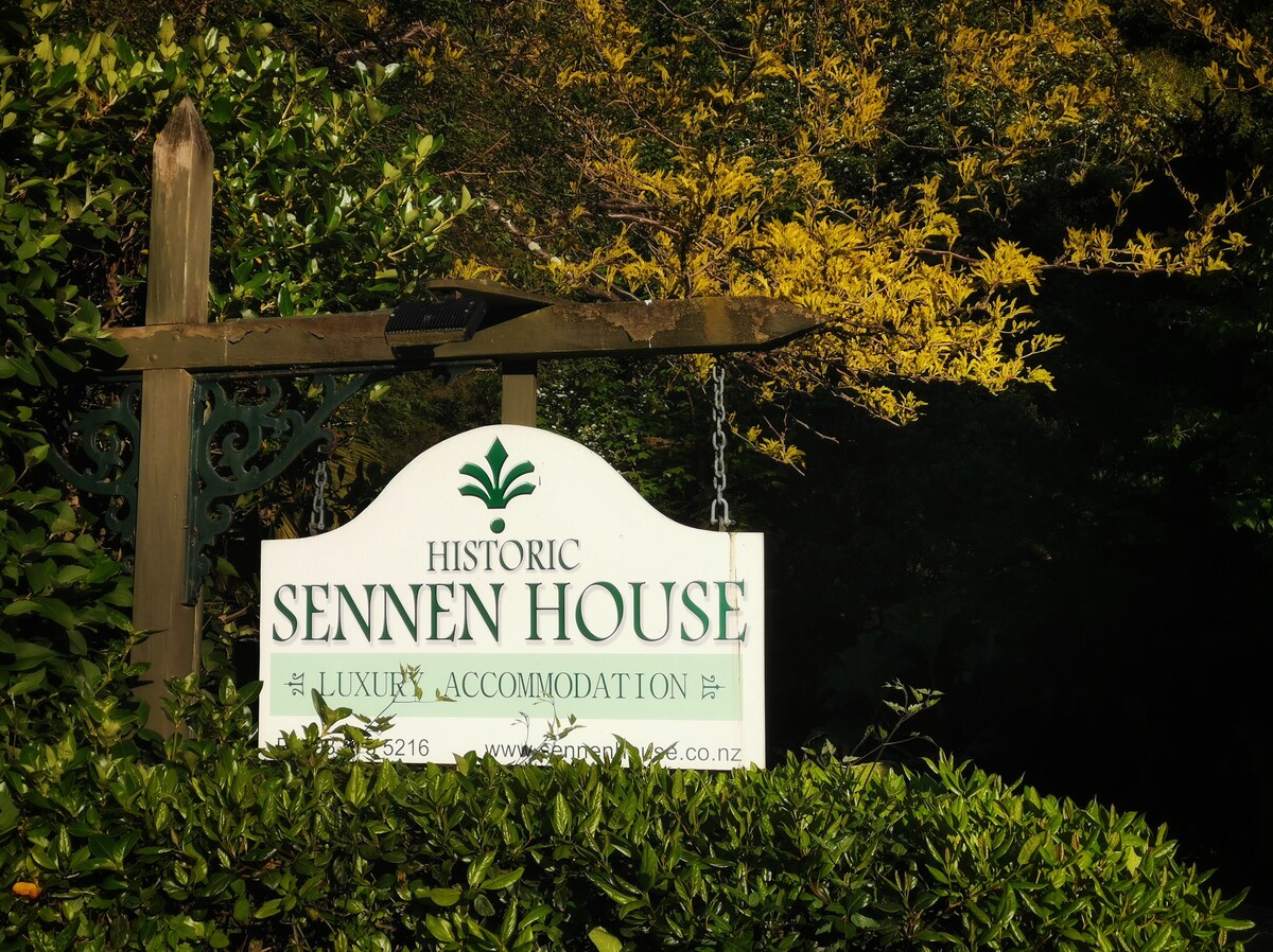 A welcoming sign for Sennen House is prominently displayed, surrounded by lush green foliage. The sign highlights the historic and luxurious accommodation, with the text clearly visible against a backdrop of mature trees and vibrant yellow flowers.