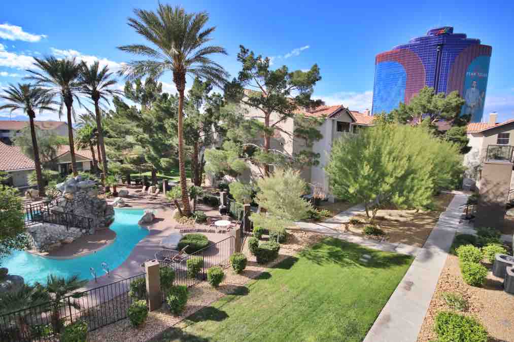 The image features a serene pool area surrounded by palm trees and lush landscaping. A rocky waterfall adds a natural element to the space, while the towering blue structure of the Rio Casino is visible in the background, contrasting with the greenery.