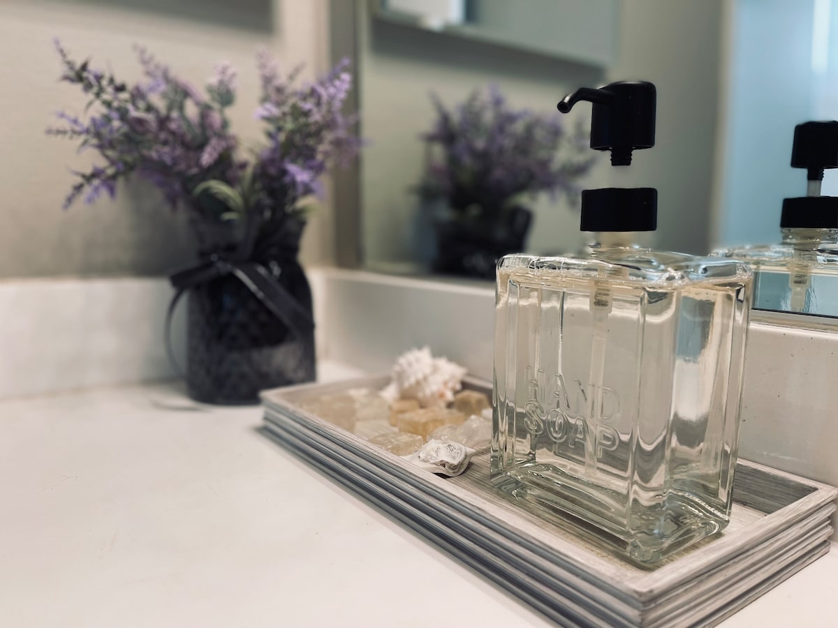 A modern bathroom countertop features a stylish hand soap dispenser and a decorative tray with seashells. A small vase with lavender flowers adds a touch of color, reflecting the clean and fresh ambiance created by the neutral tones and sleek finishes.