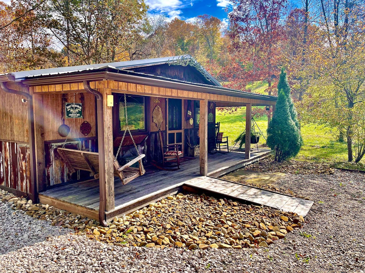The charming cabin exterior is framed by lush greenery and colorful foliage. A covered porch features hanging swings and rocking chairs, inviting relaxation. A wooden walkway leads to the cabin entrance, enhancing the rustic appeal amidst the natural surroundings.