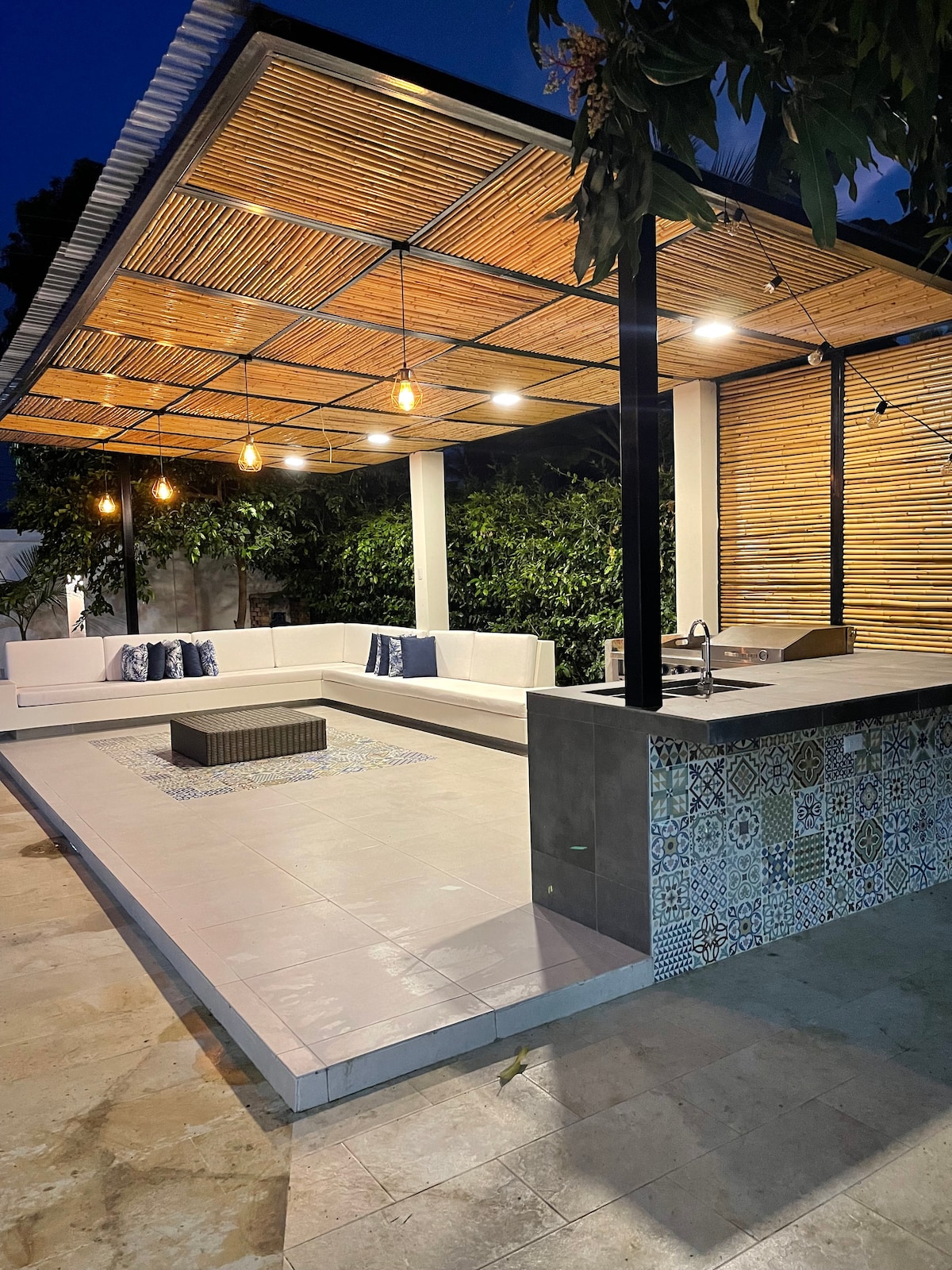An outdoor recreational area features a spacious covered seating arrangement with white cushions and decorative pillows. Pendant lights hang from a bamboo ceiling, illuminating the space. A stylish bar area is visible in the corner, showcasing colorful tiles.