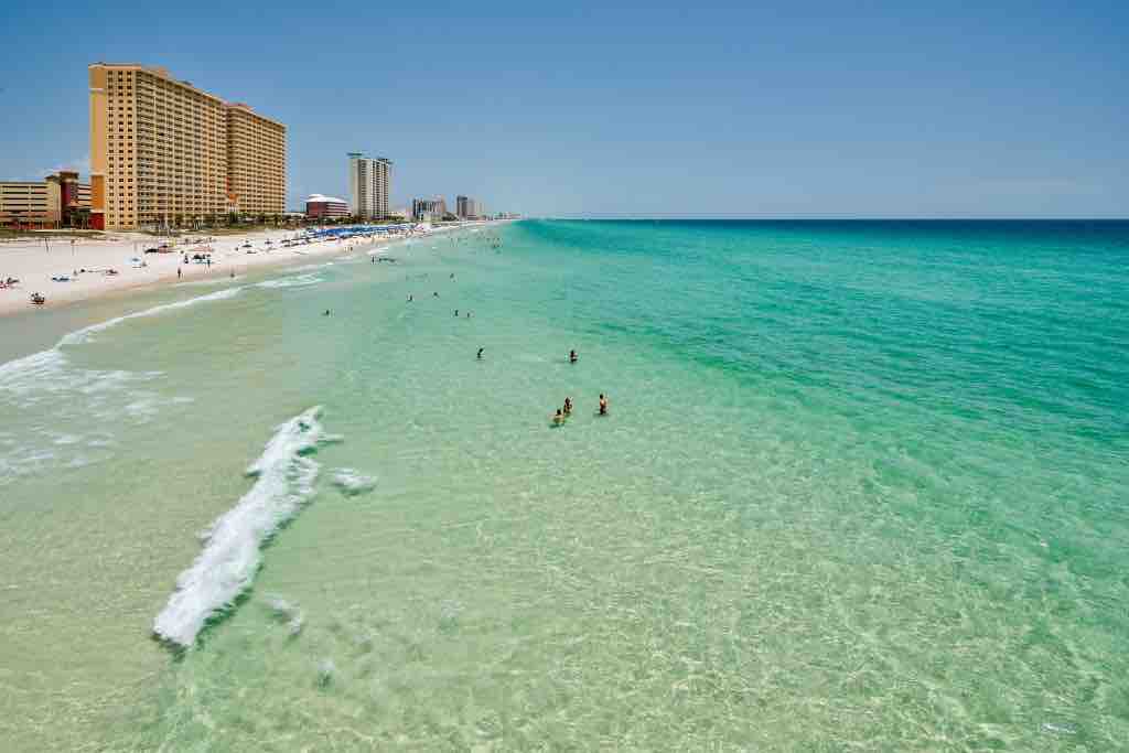 A clear view of the Emerald Coast showcases tranquil turquoise waters gently lapping against a sandy beach. Beachgoers can be seen enjoying the water, while several beachfront high-rise buildings line the shore in the background under a bright blue sky.