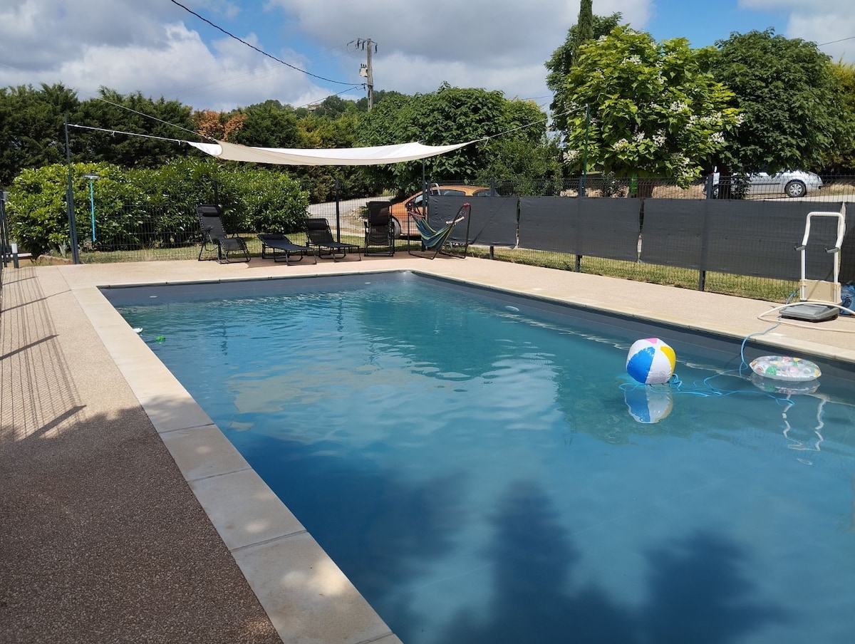 A private pool area features a clear blue water pool surrounded by a paved deck. Comfortable loungers are arranged nearby, and a sunshade provides relief from sunlight. A vibrant beach ball floats in the water, set against a backdrop of greenery and a secure fence.