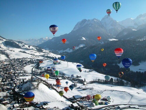 A wide view showcases numerous colorful hot air balloons floating above a snowy landscape. Rolling mountains serve as a backdrop, while a small village is nestled below, surrounded by white fields and patches of evergreen trees.