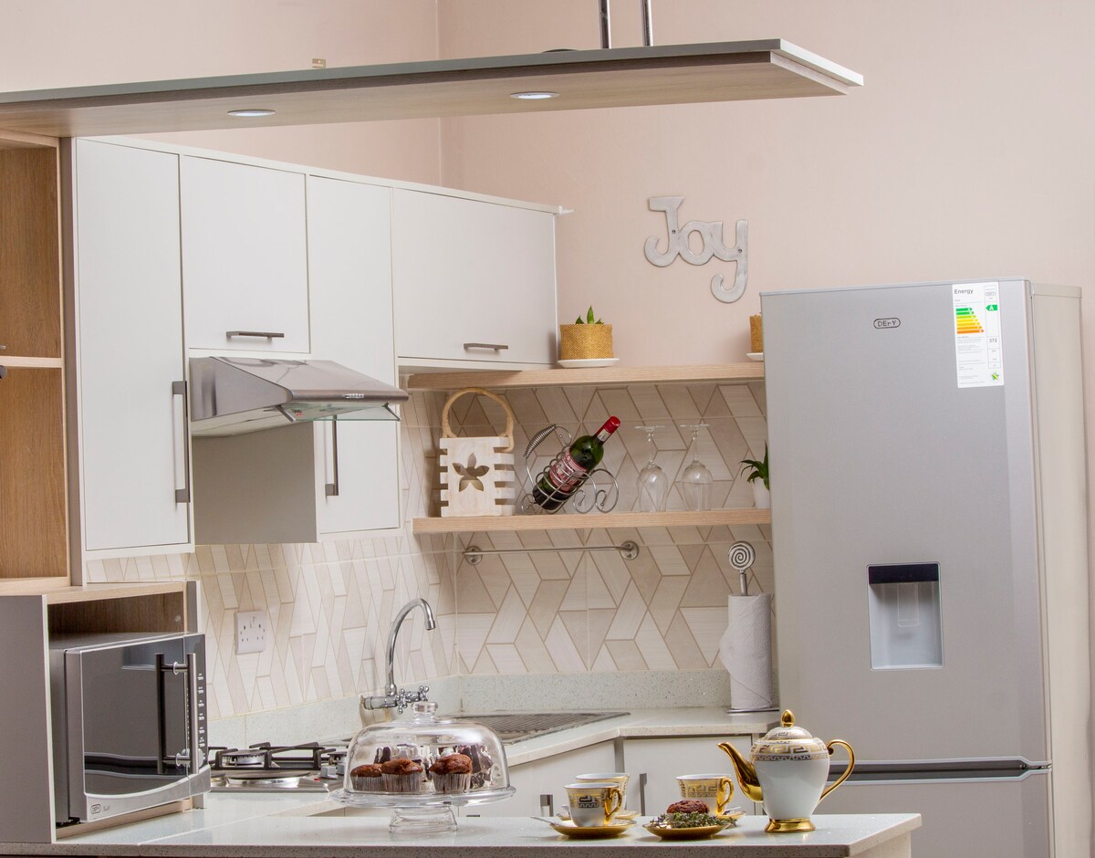 A modern kitchen features sleek cabinetry and open shelving. A stainless-steel refrigerator and microwave are included. Cooking utensils are neatly arranged, and a glass cake stand showcases baked goods, accompanied by a set of teacups and a teapot on a white countertop.