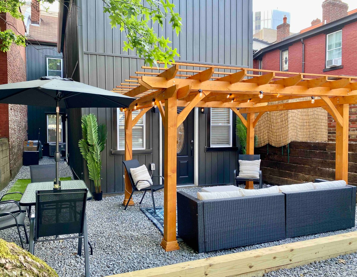 An outdoor space features a wooden pergola with soft lighting, complemented by a seating area with dark wicker furniture. A dining set and additional seating are situated on gravel, alongside a shaded area under a large umbrella, creating an inviting atmosphere for relaxation.
