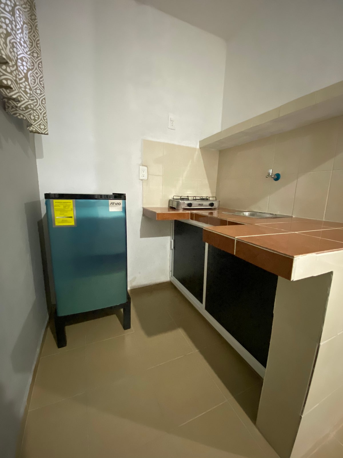 A compact kitchen area features a small refrigerator and a gas stove with two burners. The countertop is made of tiles, providing a functional workspace. Light filters in through a window with patterned curtains, illuminating the neutral-toned walls.