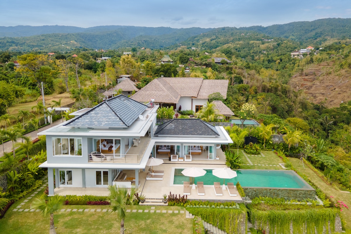 An aerial view of Villa IzaJay captures the modern architectural design and lush surroundings. The villa features multiple levels with expansive glass windows and balconies. An inviting infinity pool is visible, surrounded by lounge chairs, meticulously landscaped gardens, and a backdrop of rolling hills and tropical trees.