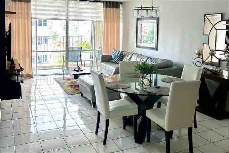 A bright and airy living area features a glass dining table surrounded by four elegant chairs. A comfortable sofa is positioned nearby, complemented by a colorful area rug. Large windows allow natural light to fill the space, showcasing views of the outdoors.