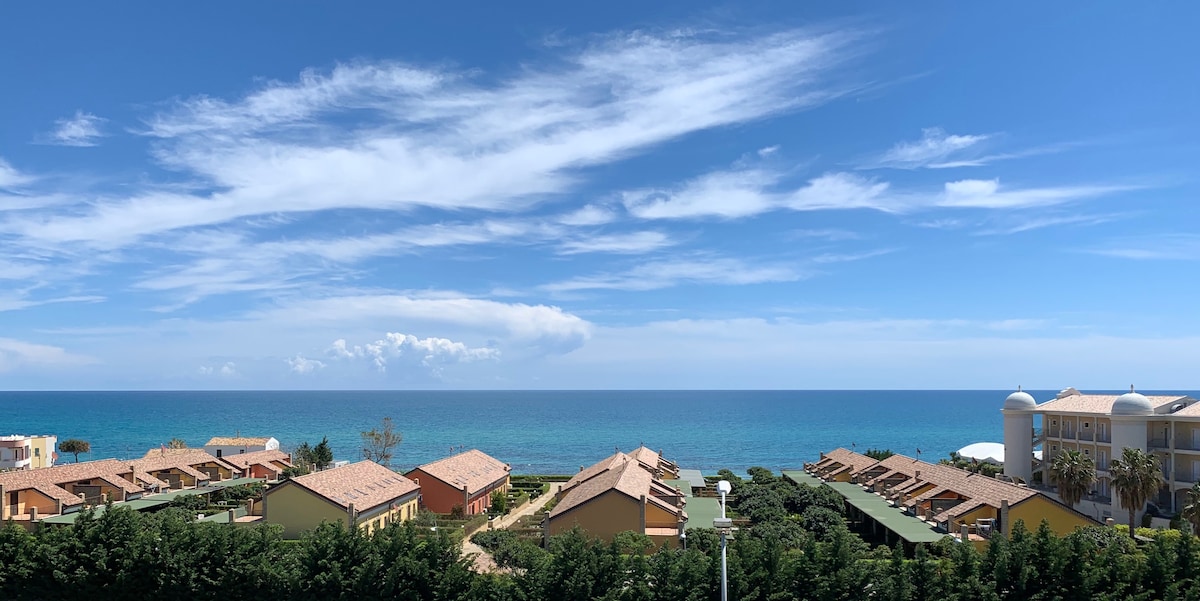 A panoramic view showcases the serene blue sea, with low-rise buildings lining the shoreline. Lush greenery is visible in the foreground, adding to the coastal scenery. The sky features scattered clouds, creating a calming backdrop for this seaside landscape.